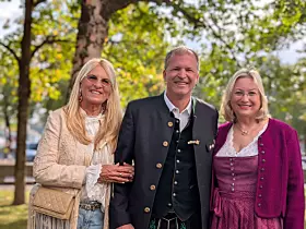 Drei Personen in traditioneller Kleidung stehen vor einem Baum und lächeln in die Kamera.