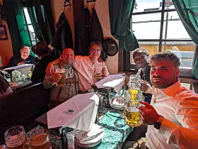 Gruppe von Gästen in einer Bar hebt Gläser an einem gedeckten Tisch mit Fenstern im Hintergrund.