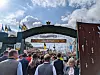 Menschenmenge unter einem begrünten Eingangsbogen beim Oktoberfest mit Fahnen und Festzelt im Hintergrund.