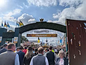Menschenmenge unter einem begrünten Eingangsbogen beim Oktoberfest mit Fahnen und Festzelt im Hintergrund.