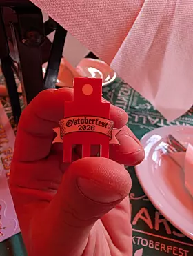 Rote Hand hält eine kleine Figur mit „Oktoberfest 2026“ vor einem gedeckten Tisch.