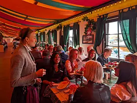 Menschen sitzen an Tischen in einem bunt dekorierten Raum und essen und trinken gemeinsam.