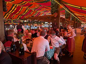 Belebtes Festzelt mit Menschen an Tischen unter einem rot-gelb gestreiften Zeltdach.