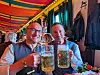 Zwei Männer halten in einem bunt dekorierten Lokal große Bierkrüge und lachen in die Kamera.