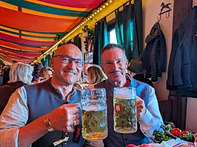 Zwei Männer halten in einem bunt dekorierten Lokal große Bierkrüge und lachen in die Kamera.
