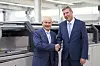 Zwei Männer schütteln sich in einer Fabrikhalle vor einer großen Industrieanlage die Hände.