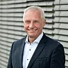 Lächelnder Mann im Anzug vor einer Fassade mit horizontalen Lamellen.