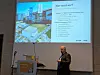 Ein Mann steht vor einer Projektionsleinwand mit einer Folie über ein Unternehmensgebäude und Stichpunkte zu „Wer sind wir?“