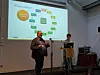 Zwei Personen vor einer Projektionswand mit Diagramm in einem Konferenzraum, eine Person spricht am Mikrofon.