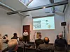 Ein Mann hält einen Vortrag vor Publikum in einem Konferenzraum mit Projektionsleinwand.