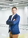 Mann im blauen Sakko steht mit verschränkten Armen in einem hellen, modernen Raum. Im Hintergrund ist eine automobile Produktionsstraße.