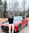 Roter Sportwagen auf einer Straße mit einem Mann im Vordergrund vor Bäumen und Bergen.