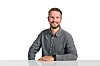 Porträt von Johannes Wörner, Produktmanager für Batteriemesstechnik bei burster. Er sitzt lächelnd an einem weißen Tisch vor einem hellen Hintergrund. Er trägt ein graues Hemd, hat kurze braune Haare und einen Bart.