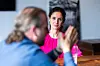 Mariella Minutolo, Vorsitzende der Geschäftsführung der ETAS GmbH, während eines persönlichen Interviews auf den ETAS Connections 2026 in Stuttgart. Sie sitzt in einer leuchtend pinkfarbenen Bluse an einem Holztisch und blickt aufmerksam zu ihrem Gesprächspartner. Ihre dunklen Haare sind zurückgebunden. Im linken Vordergrund ist der Gesprächspartner in einem blauen Oberteil stark unscharf abgebildet. Er gestikuliert mit angehobener Hand, während Minutolo ihm zuhört. Auf dem Tisch steht ein Wasserglas. Im Hintergrund sind eine helle Wand, ein angeschnittener schwarzer Bildschirm und ein abstraktes Schwarz-Weiß-Bild zu erkennen. Die Aufnahme konzentriert sich auf Minutolos Gesichtsausdruck und die persönliche Gesprächssituation.